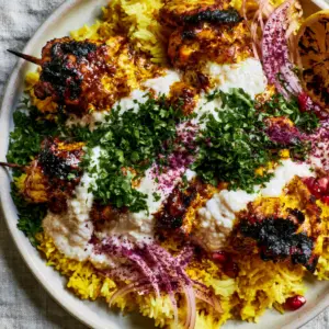 Macro shot of charred Chicken Shawarma Skewers drizzled with snowy white garlic sauce and vibrant green parsley.