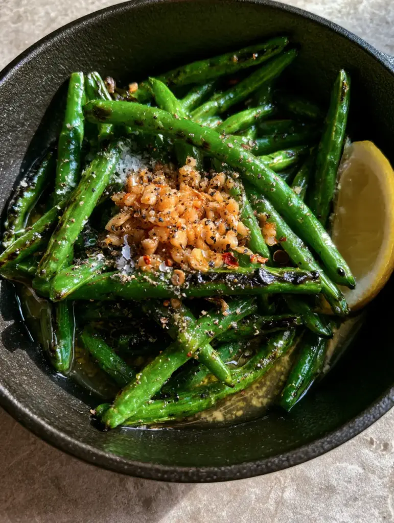 Close up of sautéed green beans topped with toasted minced garlic, flaky sea salt, and a lemon wedge.